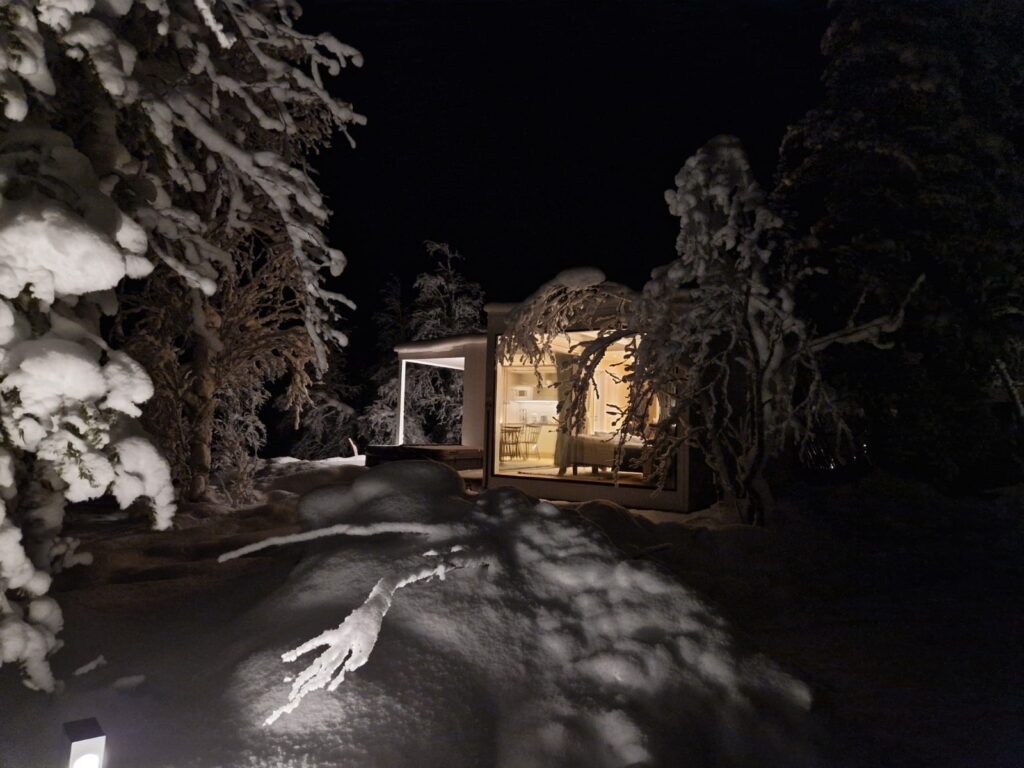 Ulkokuva Sky View Cabin Deluxe Plus -iglumökistä yöllä. Lämmin valo loistaa kinoksiin mökin suurista ikkunoista.